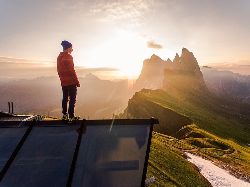 Hiker standing on mountain platform overlooking dramatic alpine peaks at sunrise with golden light illuminating jagged mountain ridges